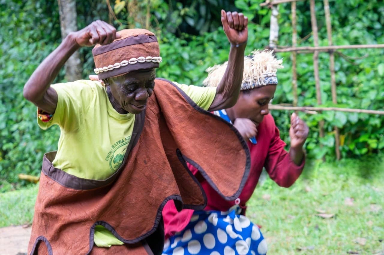 an authentic Batwa cultural dance performance in Mgahinga Gorilla National Park, Uganda, showcasing one of the most meaningful cultural experiences included in Uganda Safaris for Families: Wildlife and Culture Tours. In the photograph, Batwa men and women are dressed in traditional attire made from natural materials, moving rhythmically to ancestral songs that tell stories of forest life, survival, and identity. The lush green backdrop of Mgahinga’s volcanic landscape highlights Uganda’s rich biodiversity and cultural depth. This moment reflects how Uganda safaris for families go beyond wildlife viewing to include immersive cultural encounters that educate both adults and children. Batwa cultural dancing offers families a respectful way to learn about indigenous heritage, conservation, and community-based tourism while creating unforgettable, multi-generational safari memories in Uganda.