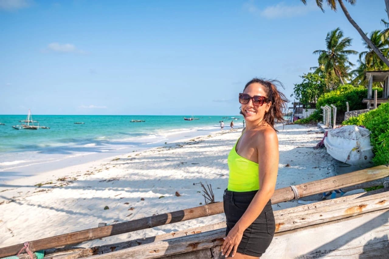 Smiling traveler standing on a white-sand Zanzibar beach beside a traditional wooden dhow at sunset — How To Visit Zanzibar: Ultimate Travel Guide