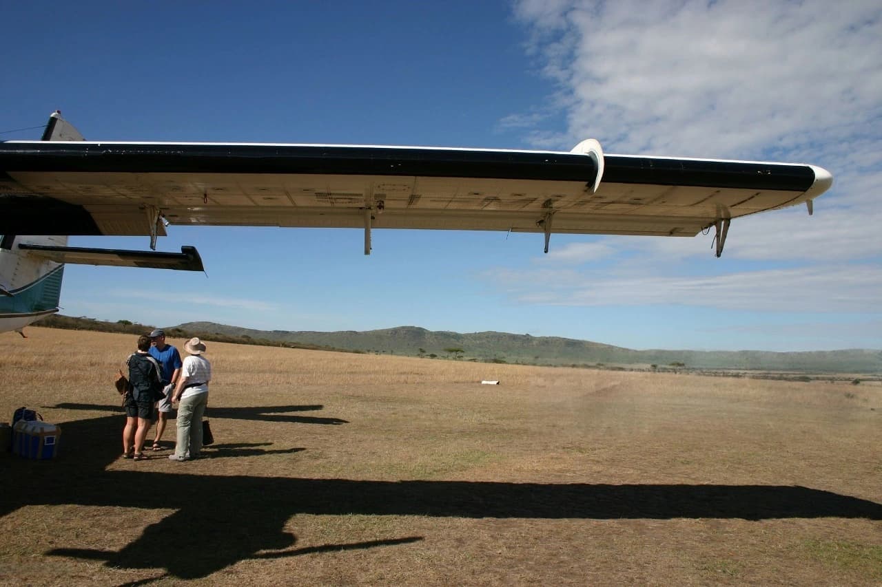 Travelers standing beside the Serengeti airstrip with their bags after arriving by small aircraft, witnessing Tanzania’s wildlife and landscapes — what to pack for Tanzania safari inspiration.