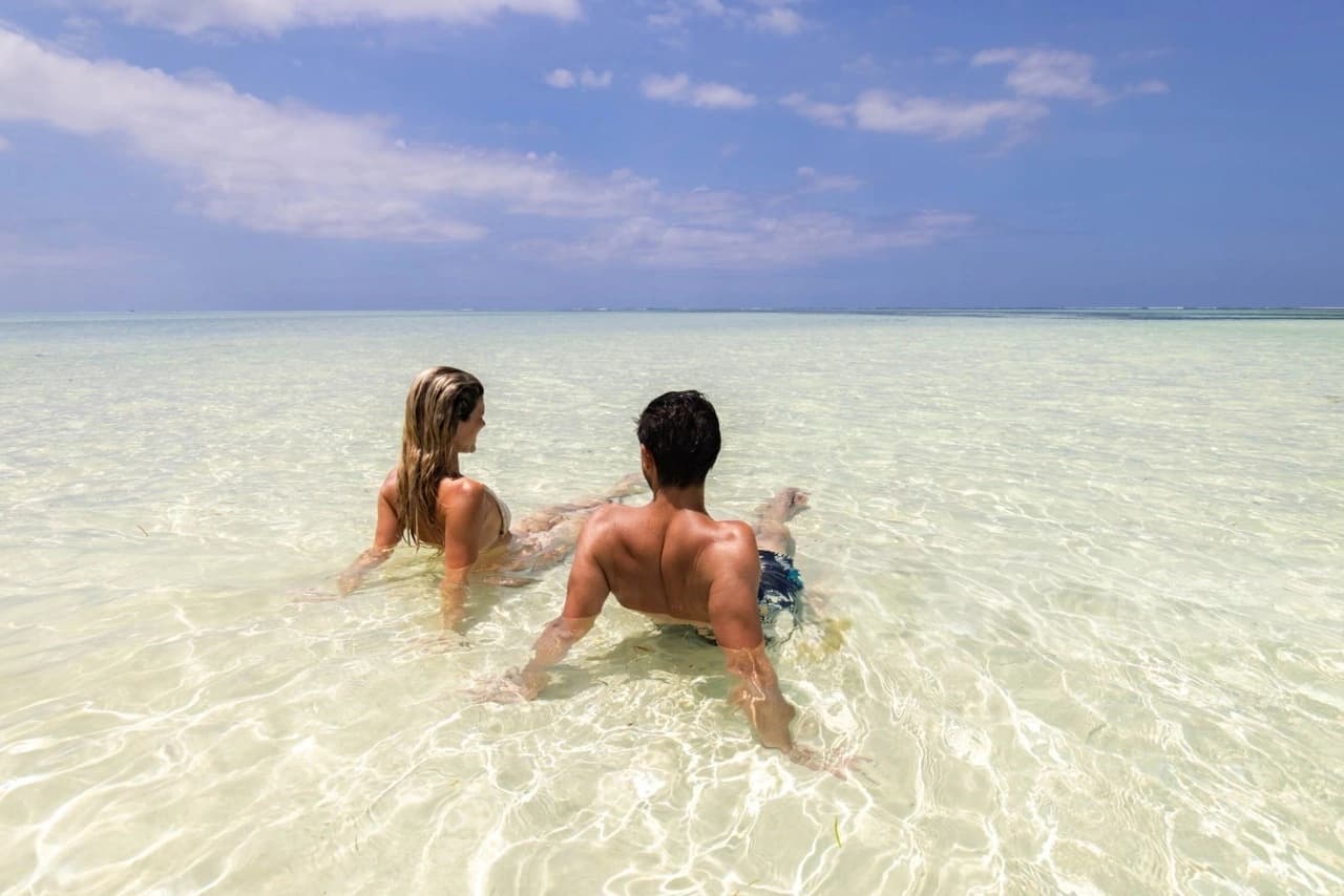 Couple on a honeymoon safari at a sun-kissed Zanzibar beach, sitting in the shallow Indian Ocean on white sand, enjoying a serene tropical landscape in true Zanzibar paradise